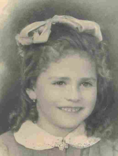 Picture of a young girl's face with long curly hair falling to her shoulders. A bow is pinned up on her head. She has an embellished collar with beads. The girl is smiling and exposes her upper teeth.
