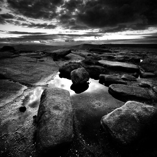 Black and white photo of rocks in the foreground, with water reflecting the light in the sky ahead.