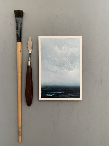 Original seascape oil painting by Tisha Mark, "Soothe Me" oil on Arches oil paper, 6"x4" (2024), shown with a paint brush and palette knife for scale. Soothing seascape painting with soft white cloud formations over a dark blue sea. Painted in a limited palette of blues and whites.