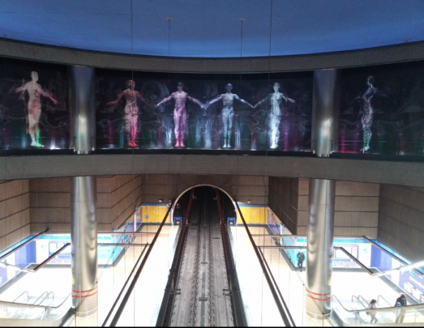 Photo of the metro station from the perspective of above looking down at the platforms and the line in the middle. There is a huge mural of 12 human figures, although only 6 can be seen as two pillars obscure the view of 2 and a further 2 are outside the shot as the mural spans so much space