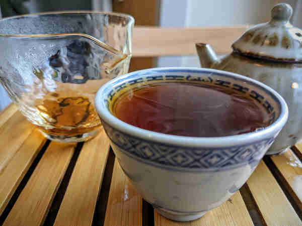 A rice grain pattern blue and white porcelain cup filled with oolong tea; a faint white mist swirls on the surface of the tea. A glass pitcher and teapot in the background.