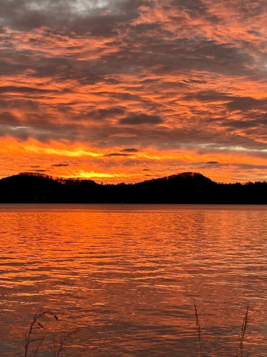 A vibrant orange sunset over a lake with silhouettes of hills in the background. The sky is filled with dramatic, textured clouds reflecting on the water's surface.