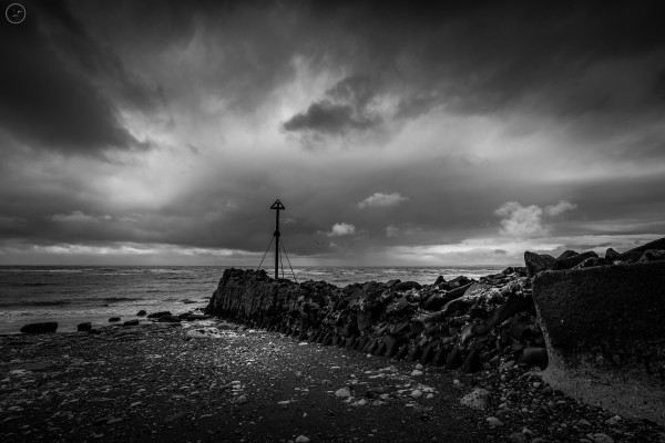 Monochrome shot of stormy skies over pebble beach with concrete break and metal warning pole