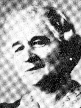 This is a black and white photograph of a woman, smiling slightly toward the left. Her hair is styled short and she is wearing a light-colored blouse. 