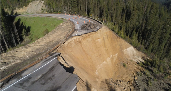 Image of a road, where most of it is missing, having been swept to the bottom of a hill by a big landslide.  Beautiful green pines are all around, except where they have been destroyed by a massive landslide... brown dirt inside the landslide