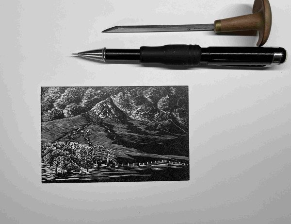 Wood engraving print in black ink. A small, rounded mountain with a rocky summit, billowing clouds behind, lit by a low sun from the left, deep shadows on the right. Pencil and engraving tool for scale.