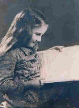 A young girl with long hair is sitting and holding a large open book.