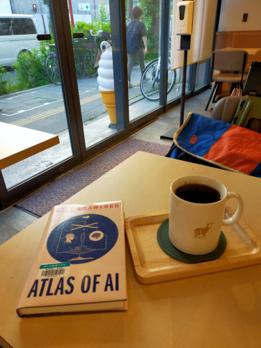 Photo is of inside a cafe. On the light tan wooden table on which is the hardcover paperback book with a blue circle with an illustration of a phrenology head looking at a tree above which are marionette sticks below which are 2 men across from each other on a grid the middle of which is a box. To the right is a white coffee cup of black coffee with a little hippo emblem on it. The cup is on a round green coaster in a small rectangular tan wooden tray. There is a blue/orange bag in the chair across the table. Outside the cafe is a large plastic soft serve ice cream cone light, to the right is a bicycle and the back of a man walking to the right