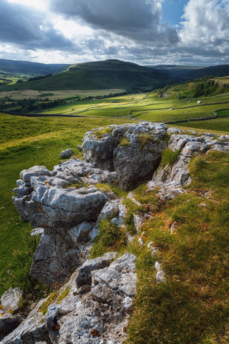 A sweeping view of the surrounding landscape. In the foreground, rugged limestone rocks are visible, their surfaces worn and weathered, interspersed with patches of grass and small plants clinging to the crevices. The midground features gently rolling green fields, divided by traditional dry stone walls that create a patchwork effect across the landscape. These walls lead the eye towards the background, where larger hills rise, covered in a mix of green pastures and darker wooded areas. The sky above is a mix of blue and clouds, casting a dynamic range of light and shadows across the scene, highlighting the undulating terrain.
