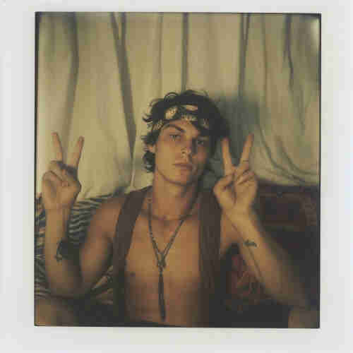 A Polaroid-style photo of a young man with dark hair. He is wearing a headband with a leaf pattern, and his chest is partially bare beneath what appears to be a draped garment or shawl. He has a necklace around his neck and a couple of tattoos on his arms. He’s giving a peace sign with both hands, has a relaxed demeanor, and appears to be sitting against a backdrop of a light-colored curtain with a patterned pillow or textile behind him. The setting evokes a bohemian or retro vibe, reminiscent of the 1960s or 1970s counterculture aesthetic.