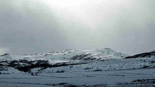 An almost monochrome photo of a snow scene. The foreground is in partial shadow as the sky is almost completely overcast. There are dark trees in a valley, lending scale to the shot. On the horizon a peak of ice and black rock is highlighted by sunbeams fanning out softly above the mountain top.