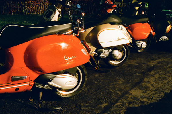 Three Vespa moto scooters seen one behind the other. The first and third are red, the second is white. The surroundings appear dark. olour photo.