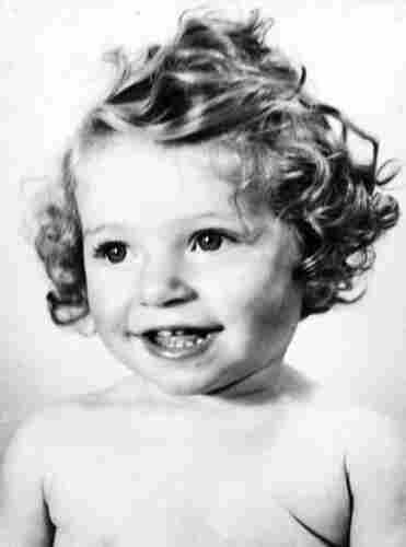 A photo of the face and shoulders of a baby girl. She is smiling. She has long, curly hair. 