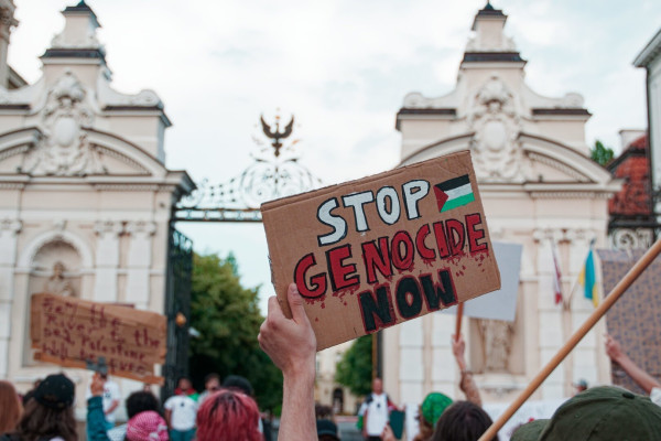 Someone's hand holding a carton board saying "Stop Genocide Now" in front of the main gate of the University of Warsaw. Blurry crowd and more boards can be seen nearby.