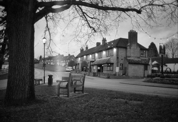 Black and white image shows a bench, facing away, under a tree. Behind to its right is a building with lights on it what is clearly early twilight (this is the Queen and Castle pub/restaurant). Behind the bench to the left is a lit street lamp, and a main road curving up the hill. On roughly the left hand third a vertical light leak is reminiscent of the transporter beams from sci-fi movies!