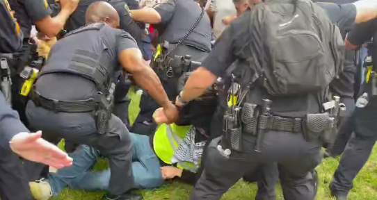 A young person in a hi-viz and keffiyeh is wrestled and pinned to the ground by several armed police.