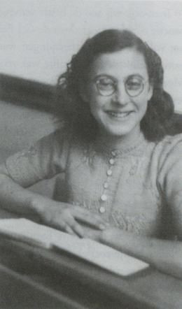 A historical black-and-white photograph of a Dutch Jewish girl Suze Alexander, smiling while seated at a desk with an open book in front of her. She is wearing round glasses and a buttoned-up blouse.