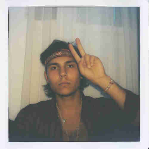 A photograph of a young man in a Polaroid-style frame. He’s wearing a bandana around his head and has medium-length hair. The man is giving a peace sign with his fingers and is dressed in a buttoned-up shirt with a dark, plaid pattern. He has a necklace and a bracelet, which add to his bohemian look. The background appears to be a room with white curtains, and the photo has a soft, vintage feel to it, reminiscent of the 1970s era.