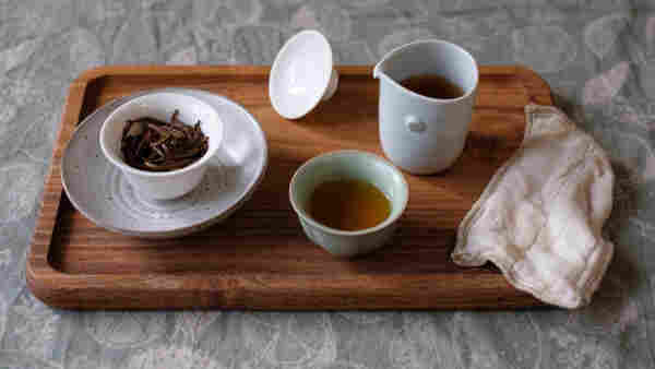 wooden tea tray with small porcelain gaiwan showing black tea leaves, in a ceramic cup, pitcher, green porcelain cup