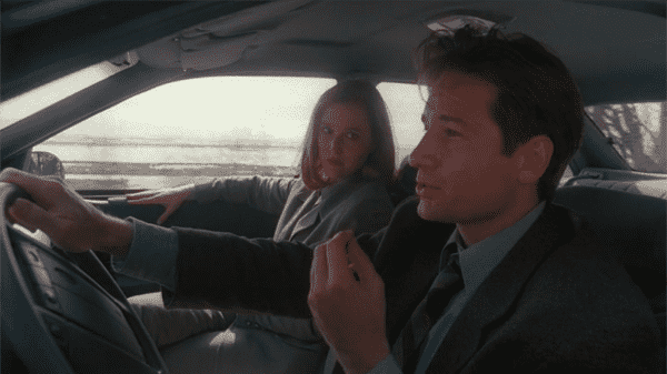 A man and woman drive in a car. The man speaks to the woman, who looks skeptical