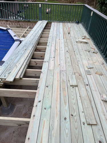 View of an outdoor deck under construction.  Image shows several rows of top boards that have been installed as well as open joists and a brief view of a round pool.