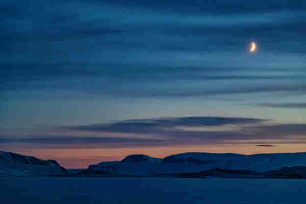 A crescent moon hangs in the sky. Dark blue, purple, and grey clouds run across the sky. The sky itself is the colour of denim, fading as it gets closer to the horizon, but near the horizon the colour changes to purple, pink, and orange.  The sea ice and mountains are bathed in darker blues. 