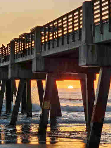 Standing beneath a massive pier extending into the surf of a sandy beach at sunrise with the sky filled with shades of yellow and orange above the crashing waves and splashing of water against the wood pier and wet beach sand.