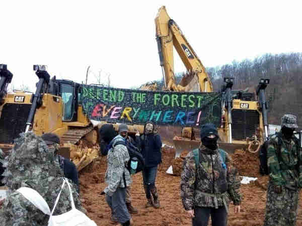Masked protesters stop pipeline construction. Banner reads, "Defend the Forest Everywhere!" Placed against construction equipment. 