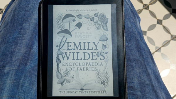 A e-reader on my lap, displaying the cover of the book Emily Wilde's Encyclopedia of Faeries by Heather Fawcett. Cover design is  all caps title in an old fashioned typeface, framed by illustrated mushrooms, ferns, and vines, and an open book exuding sparkles. Also states "Sunday Times Best Seller" 