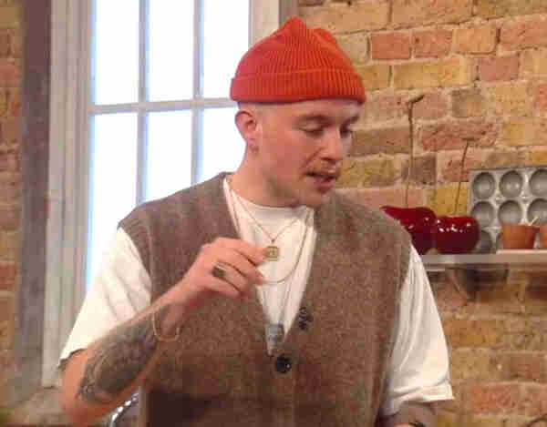 Young fellow. He's got the works. Sleeve tats. He's got a big gold chain. He's got a tweed waistcoat over a white tshirt. He's got an ironic moustache. And of course the ubiquitous orange beanie hat, worn way too high up over a shaved head. Probably concealing some terribly ironic hairstyle. He's in a kitchen. Talking about vegan food, probably using slang words. God I hate young people. 