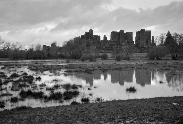 The silhouette of a ruined castle is reflected in the still waters of a flooded meadow. The left foreground has bunches of reeds poking through the water, indicating this floods quite often. Raggedy clouds above, more rain to come. Black and white photo.