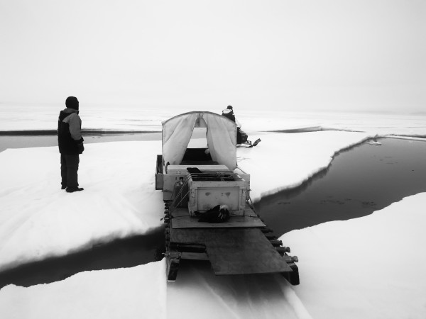 Greyscale photo. A skidoo pulling a qamutiq (sled) with an iglutak (hit) and grub box. The skidoo is on a large pan of ice that is bridging a wide crack in the sea ice. The qamutiq is partially across the crack onto the pan. A man stands on the pan of ice watching. 