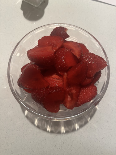 Sliced strawberries in a small glass bowl on a counter. The strawberries are deep, ripe red: vivid red all the way through; red like the lipstick color you can never find; red the way blood must look to vampires; sapsucker head red; the red of a rose cut for romance; juicy red, strawberry-red through every millimeter without an iota of compromise. 