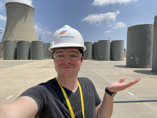 A young man standing in front of 5 m tall concrete containers with spent nuclear fuel inside stored outside, with a large cooling tower in the background