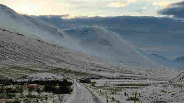 A landscape photo of a snow scene. An icy dirt road is snaking away from the viewpoint towards two impressive mountain peaks. On the valley floor there is brown scrub and small trees but the whole scene is ice- and snow-covered. The sky is overcast by a thick blanket of cloud but with pale cyan sky visible above. The curved mountains in the centre of the view are surrounded by loose snow that has been lifted by the winds and is suspended in clouds, making the hard ice look velvety and soft.