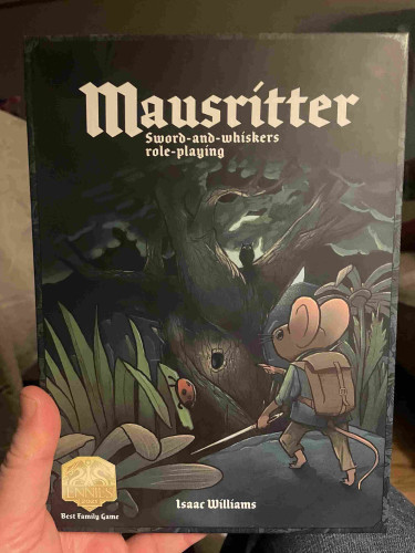 A hand holding a box set titled "Mausritter Sword-and-whiskers role-playing" by Isaac Williams, featuring illustrations of adventuring mice, with an award sticker for 'Best Family Game ENnies 2021'.