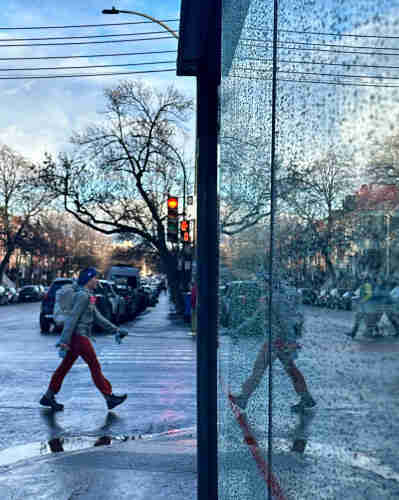 Street  scene, person walking reflected in water-speckled glass of bus shelter.