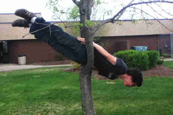 Photo of a youngish person “planking” (holding their body in a stiff, straight position) in a fork in a tree trunk, head pointing towards the ground at an angle of about 30°. It’s a metaphor for the lunar lander, you see…