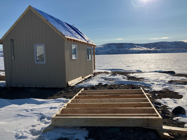 A floor framed out in preparation for an addition on the cabin. The cabin stands behind the new joist. A bright blue sky on a sun shining day. Tundra is peaking through the snow. 