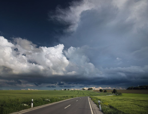 Photo of a random road, winding. At the horizon a giant thunder cloud towering like a mountain. It's lit up by the sun and casts an ominous shadow below