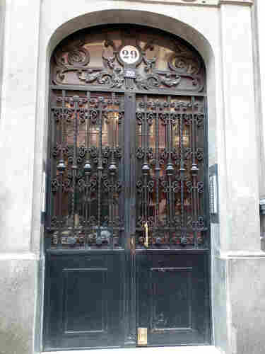 Photo of a double door that is painted black wood with glass panels in the upper half and in a panel above.
There is decorative ironwork in front of the glass panels