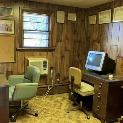 The image shows a room with a vintage aesthetic.

The room has wood-paneled walls and a patterned carpet. It contains a desk with a desktop computer, a desk chair, a green armchair, an air conditioning unit, and framed photos or certificates hanging on the wall.

The room appears to be an office, possibly in a home. The overall style suggests that the photo was taken in the 1970s or 1980s.