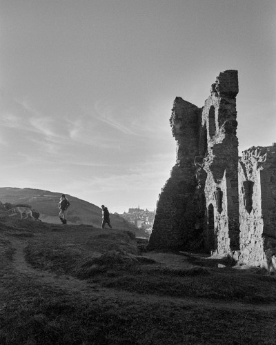 High contrast black and white portrait format photo showing part of the ruined chapel on the hill in Holyrood Park, in the afternoon sun. The foreground is mostly in shadow, with a path running through it towards two figures coming over the hill. Through a gap between the far hill and the ruin, Edinburgh Castle can just be seen on the skyline. The sky is clear except for a few wispy clouds.