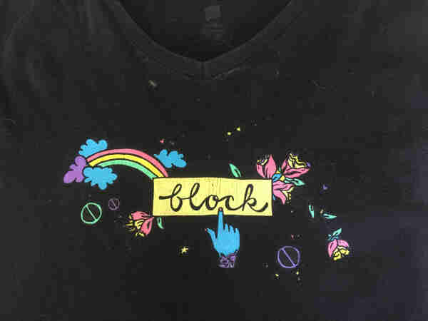 The front of a black t-shirt. There's a yellow rectangle with there word block written in it. A blue hand is pointing to the rectangle as though it's a button it's about to press. There's also a rainbow and some flowers. 