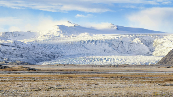 A photo of a landscape in winter. The foreground is sparse brown vegetation over gravel. In the centre of the shot is a snow-covered glacier, a fall of ice down a mountain face. Above it are two distinct peaks, spindrift snow is whipping from them showing that the wind is from the right. Some dark bare rock is visible but mainly the mountain is covered in ice. The glacier begins quite high up on the mountain, and pours in slow motion towards a ridge, where it drops suddenly before levelling out again near ground level. The sky above is cloudy but blue, and the sunlight is from the upper left. Power pylons are visible at the snout.