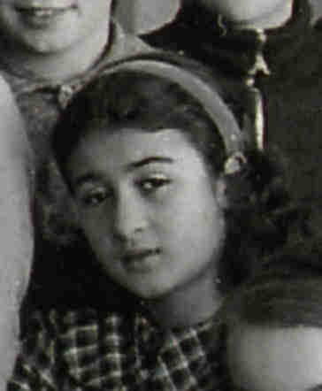 Black and white photograph of a girl with long wavy hair with a headband. Only her head and torso are visible. She is standing among other people - their silhouettes are visible all around. She is wearing a checked blouse.