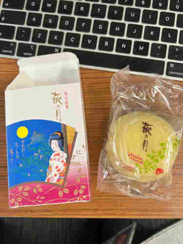 A small rectangular box with a picture of a woman in a kimono and the full moon. Next to it is the treat it contained, which was a small round yellow cake. 