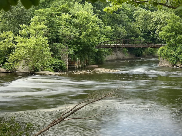 The cuyahoga river; the water is blurred from a long exposure effect. The river is flowing from the left and curves upward in the photo. There is a footbridge over the river but the end is obscured by the forest. 