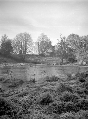 In the uper centre of this image the buildings of Kenilworth Castle are partly obscured by trees. Below it, a flooded area has frozen into an icy pond. In front of that, at the bottom of the image clumps of rough grass and other meadow plants are covered with frost. Black and white photo in portrait format.