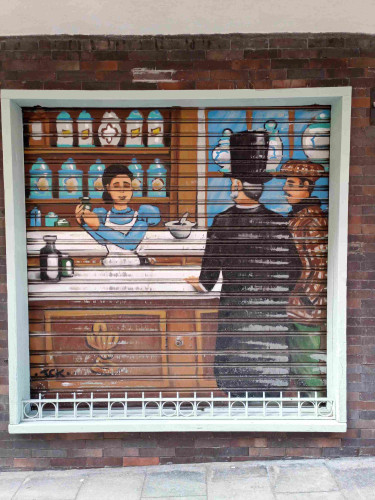 Photo of a shuttered window. The shutters have been sprayed with the scene of a lady working in a pharmacy shop serving two men in hats and coats. The paint is worn in places from where the shutters are raised and lowered